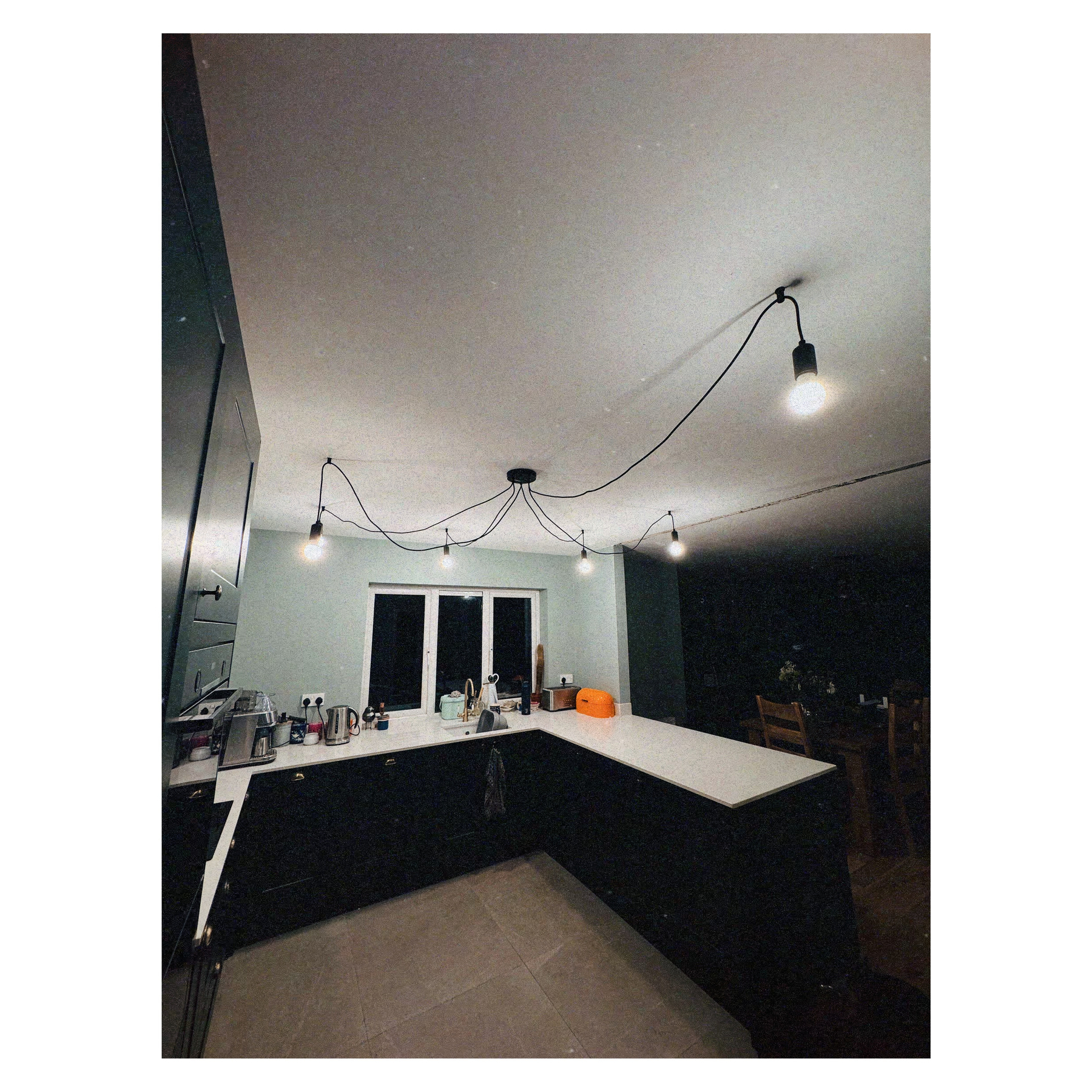 Auto-generated description: A modern kitchen features hanging pendant lights, a countertop with various kitchen items, and a dining area in the background.