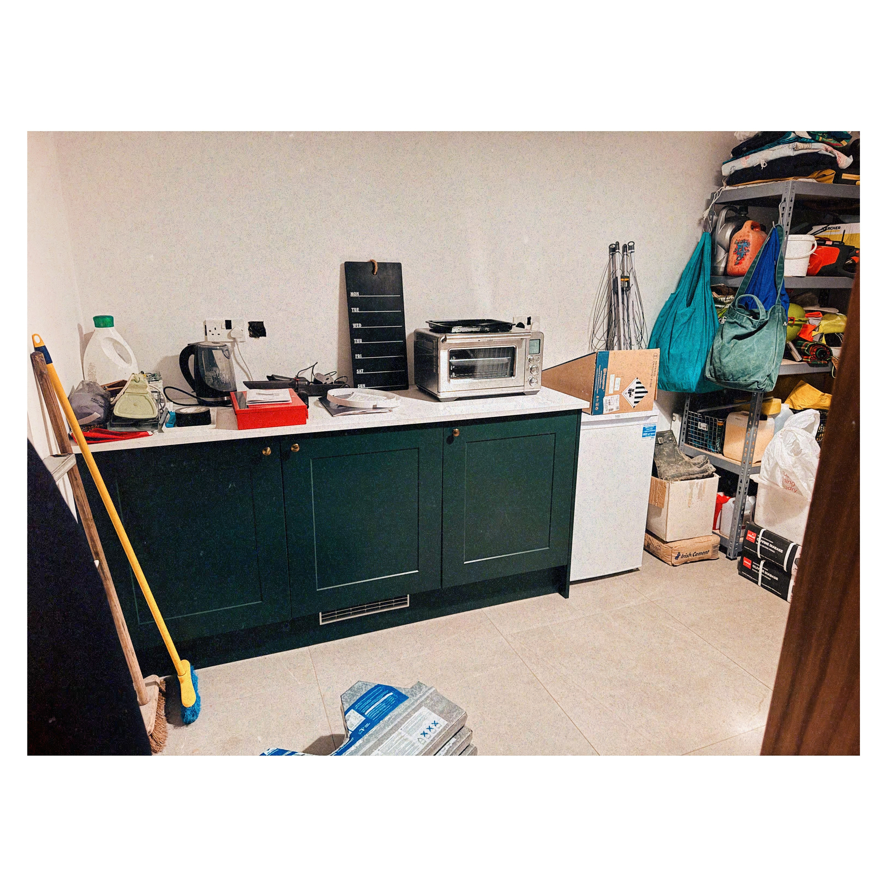 A cluttered room features a green cabinet with a countertop holding a kettle and toaster oven, surrounded by a broom, equipment, shelves, and boxes.