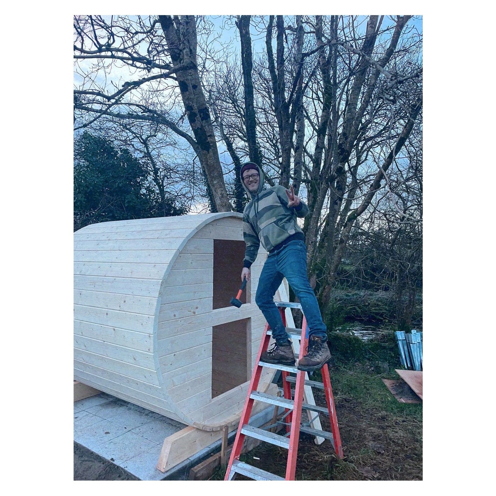 Auto-generated description: A person standing on a ladder is flashing a peace sign while working on a wooden structure in a wooded area.