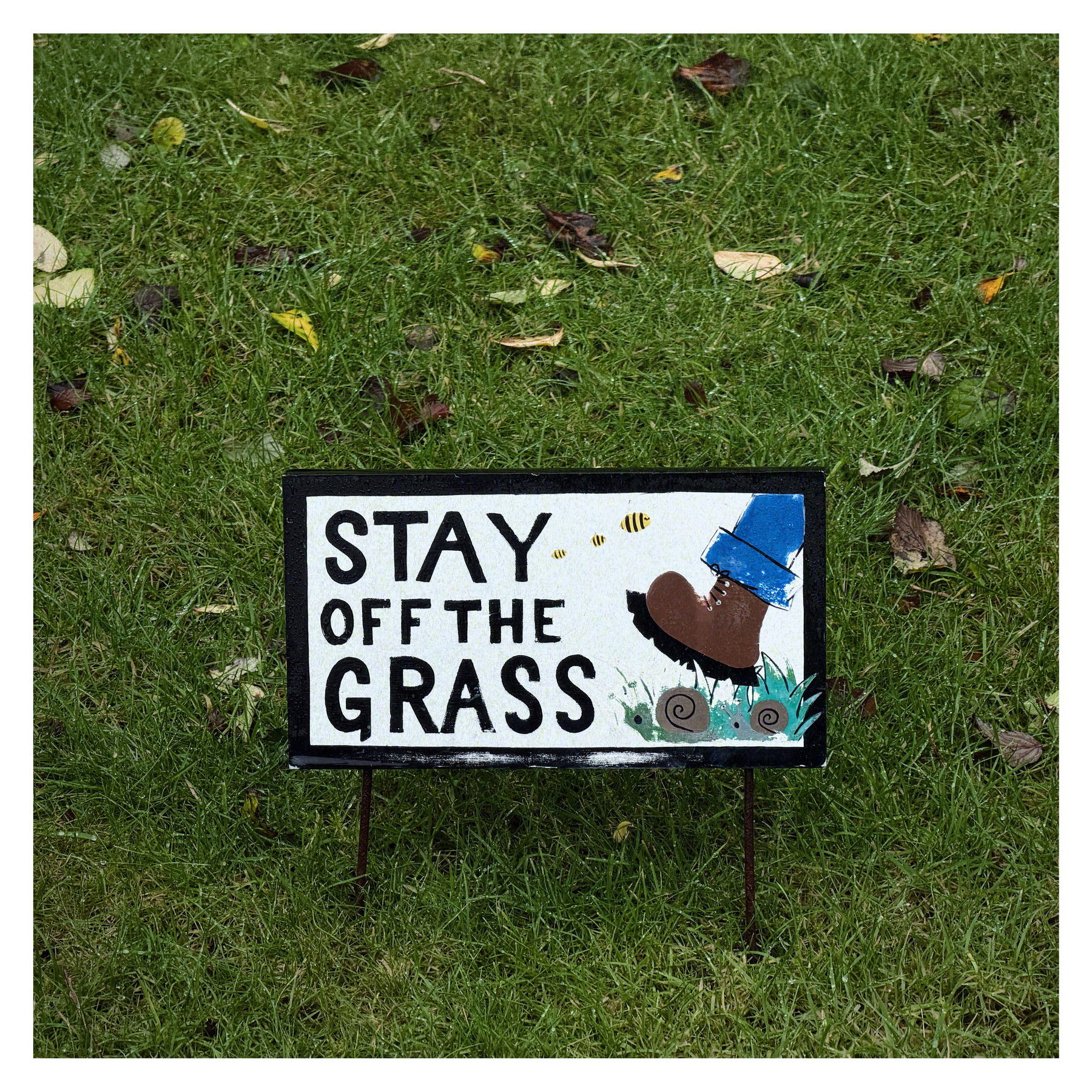 Auto-generated description: A sign on the grass warns people to stay off, featuring an illustration of a boot stepping and a bee flying nearby.