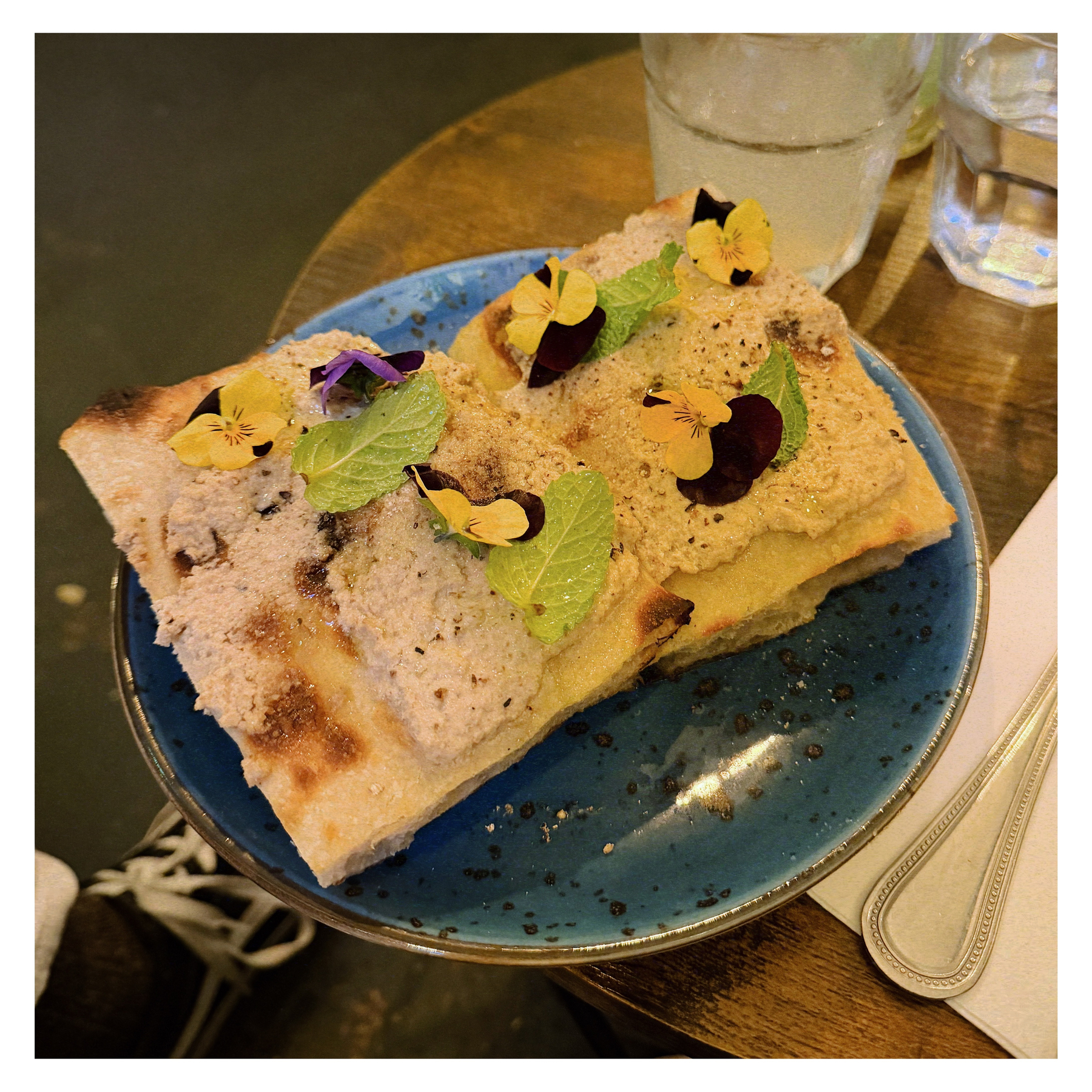 A plate of sliced focaccia topped with a spread, edible flowers, and fresh herbs is served on a blue dish.