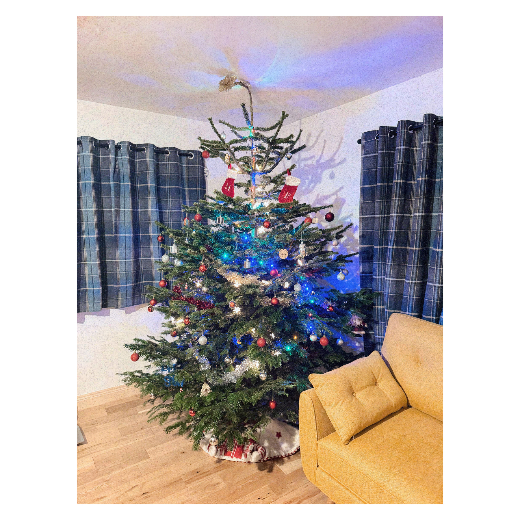 A decorated Christmas tree with colorful lights and ornaments stands next to a yellow couch in a room with checkered curtains.
