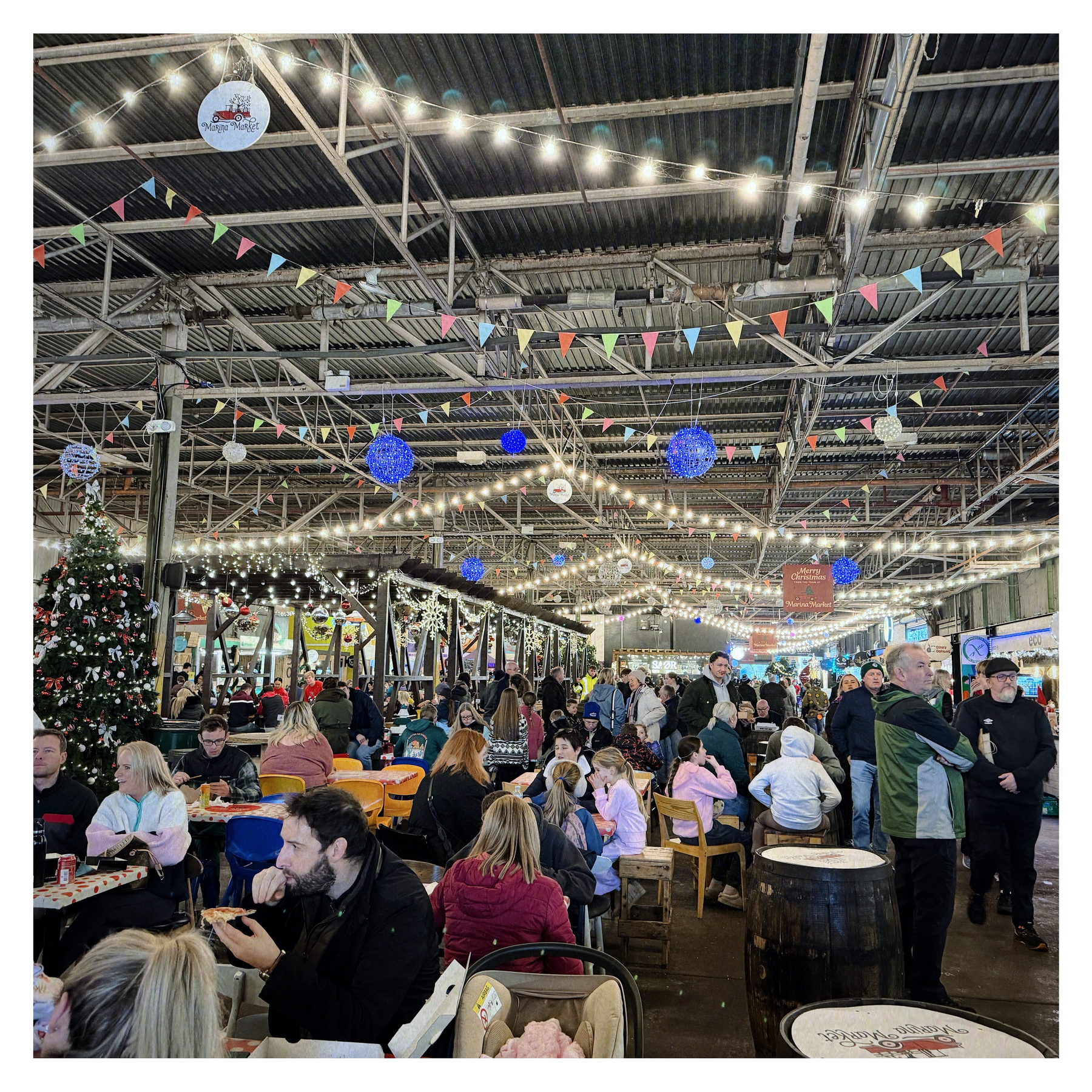 Auto-generated description: A lively indoor market scene features a festive ambiance with numerous people gathered under string lights and colorful decorations.
