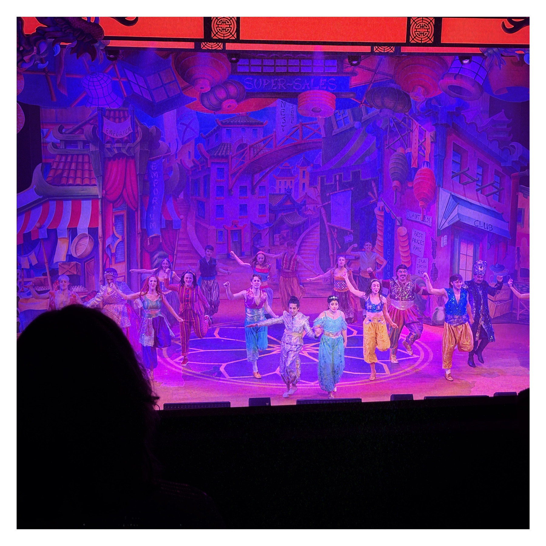 Auto-generated description: A vibrant stage performance features a group of dancers in colorful costumes against a lively, painted urban backdrop.