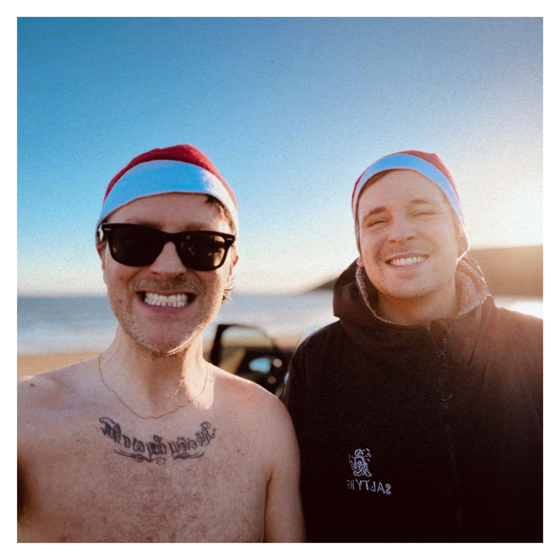 Auto-generated description: Two people wearing Santa hats, one shirtless and smiling with sunglasses, and the other in a jacket, stand on a beach with a car and the sea in the background.