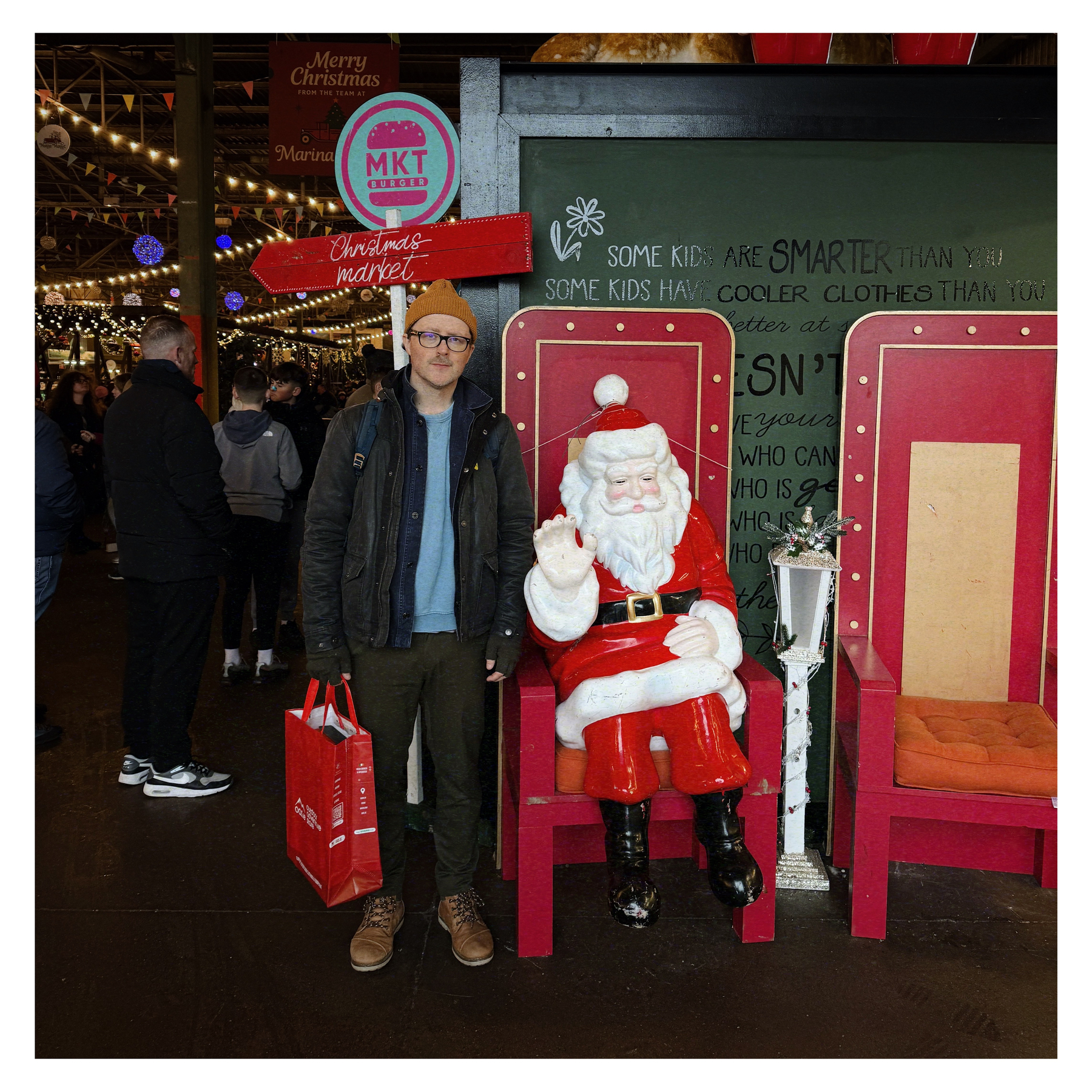 Auto-generated description: A person standing next to a Santa Claus figure in a festive market setting, holding a red shopping bag.